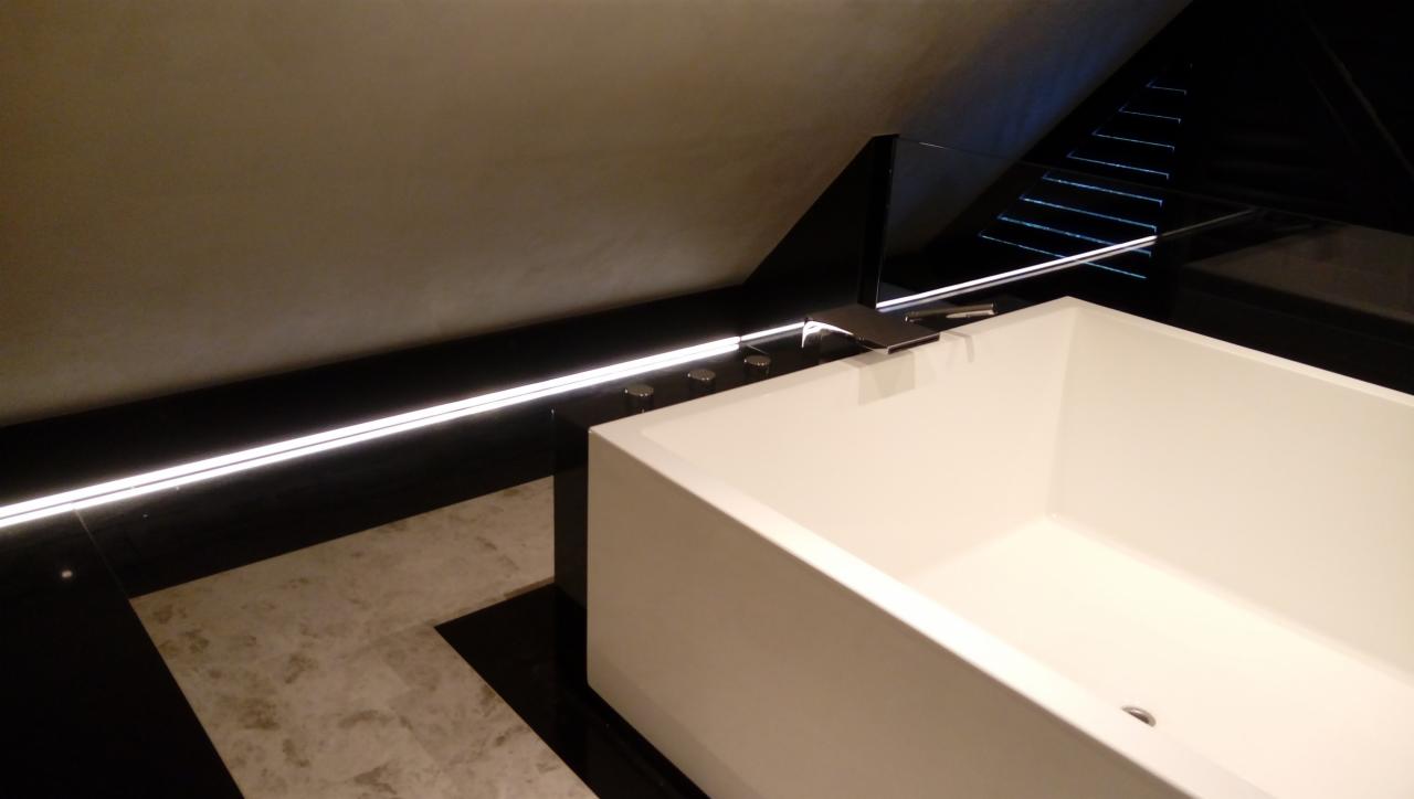 Nowoczesna biała wanna prostokątna z chromowaną armaturą, podświetlenie LED wzdłuż ściany, czarno-beżowa marmurowa posadzka w łazience na poddaszu.