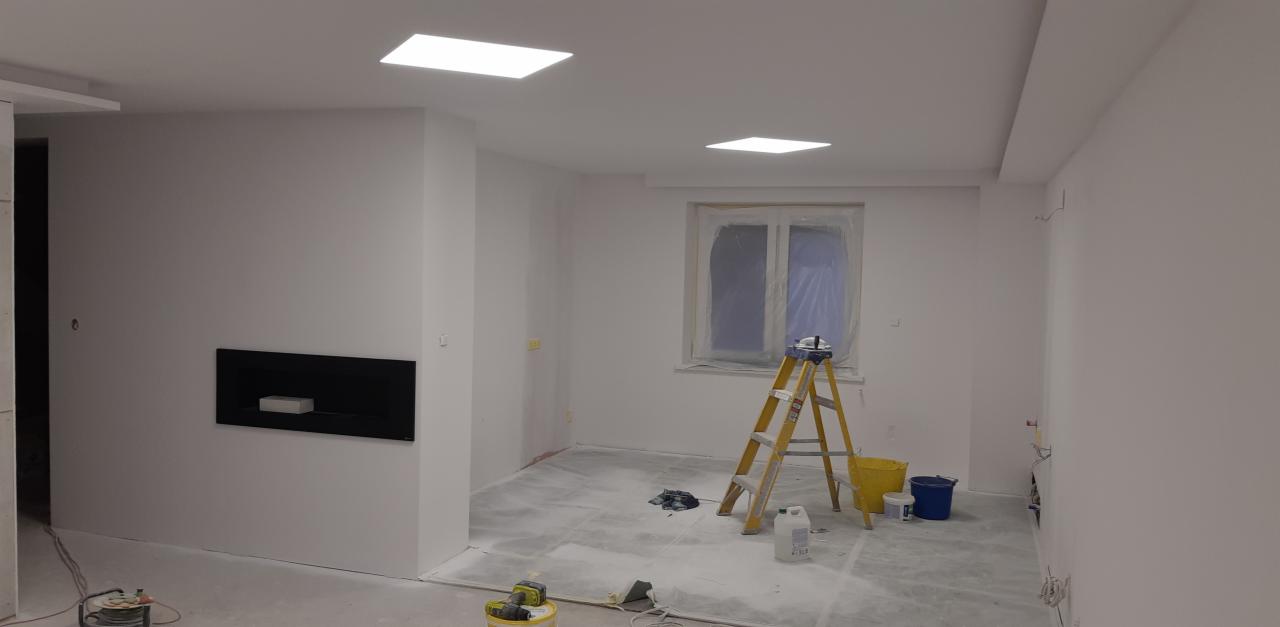 Pokój w trakcie remontu: świeżo pomalowane na biało ściany, zabezpieczone okno folią, drabina malarska, wiadro z farbą i akcesoria malarskie na podłodze pokrytej folią ochronną, czarny wkład...