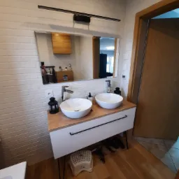 Nowoczesna łazienka z dwoma okrągłymi umywalkami nablatowymi, białą cegłą na ścianach, drewnianym blatem i czarną lampą nad lustrem, widoczne także akcesoria łazienkowe i dziecięcy stołek.