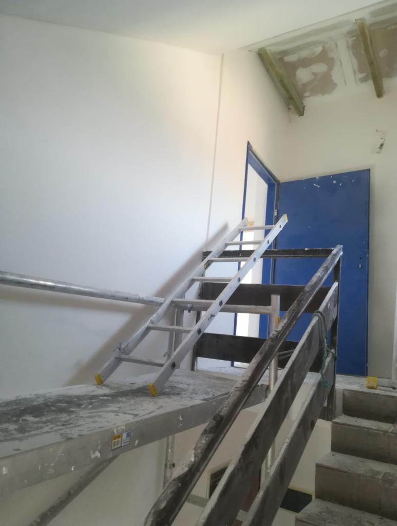 Wnętrze w trakcie malowania: rusztowanie, drabina oparta o świeżo pomalowaną ścianę, niebieskie drzwi w tle, widoczne ślady po pracach na schodach i suficie.