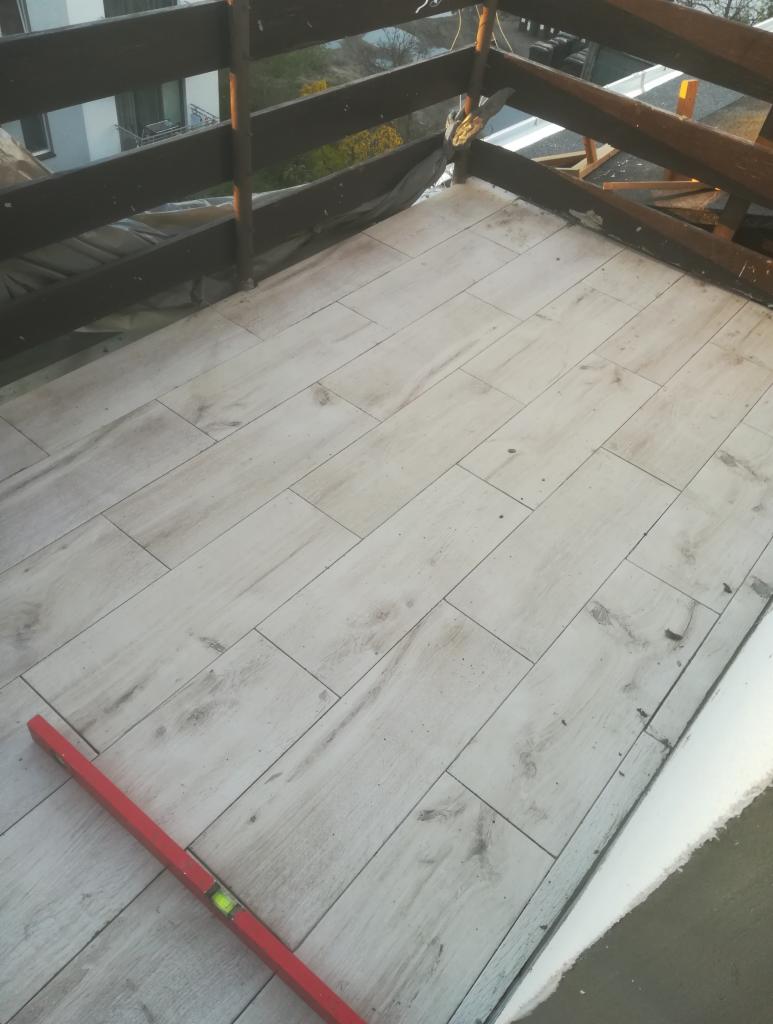 Balkon z ułożoną podłogą z płytek imitujących drewno, w tle drewniana balustrada i budynek, na podłodze leży czerwona poziomica.