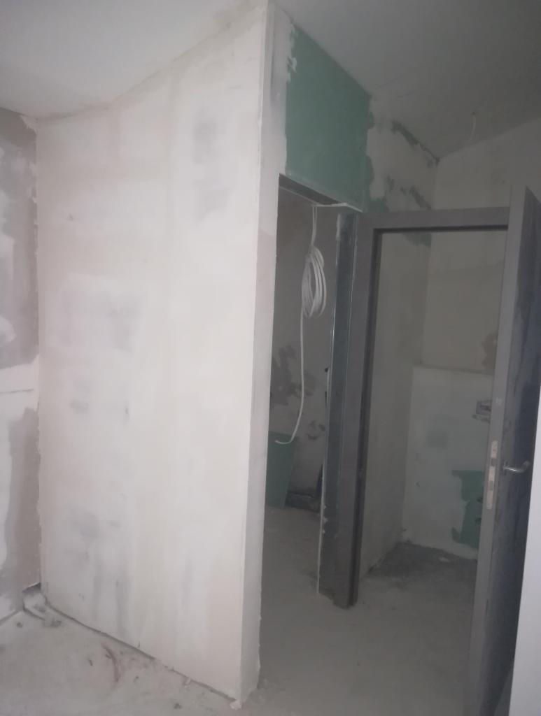 Wnętrze w trakcie prac przygotowawczych: szpachlowane ściany, widoczne przewody elektryczne zwisające z sufitu, surowa ościeżnica i częściowo otwarte drzwi.