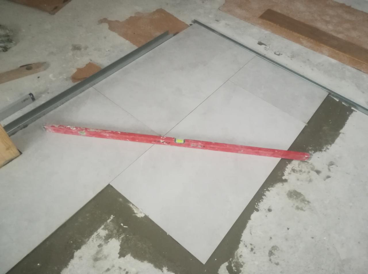 Układanie jasnych, kwadratowych płytek podłogowych z użyciem poziomicy i metalowych profili, widoczny świeży klej.