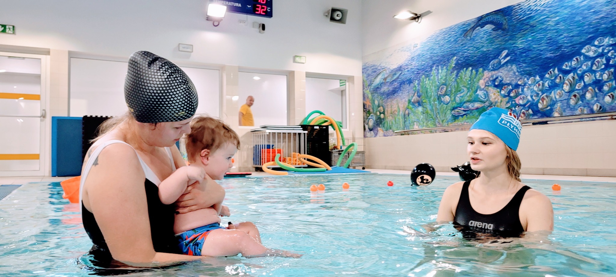 Instruktorka z dzieckiem w basenie podczas zajęć. Kobieta w czepku z dzieckiem w ramionach, druga instruktorka w wodzie, dekoracja z motywem morskim w tle.