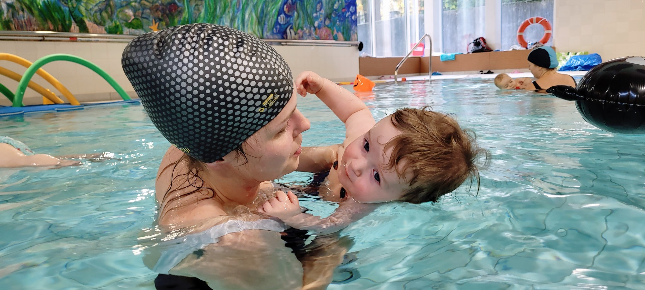 Instruktorka w czepku podtrzymuje niemowlę w basenie podczas zajęć. W tle widać inne osoby i akcesoria do pływania.