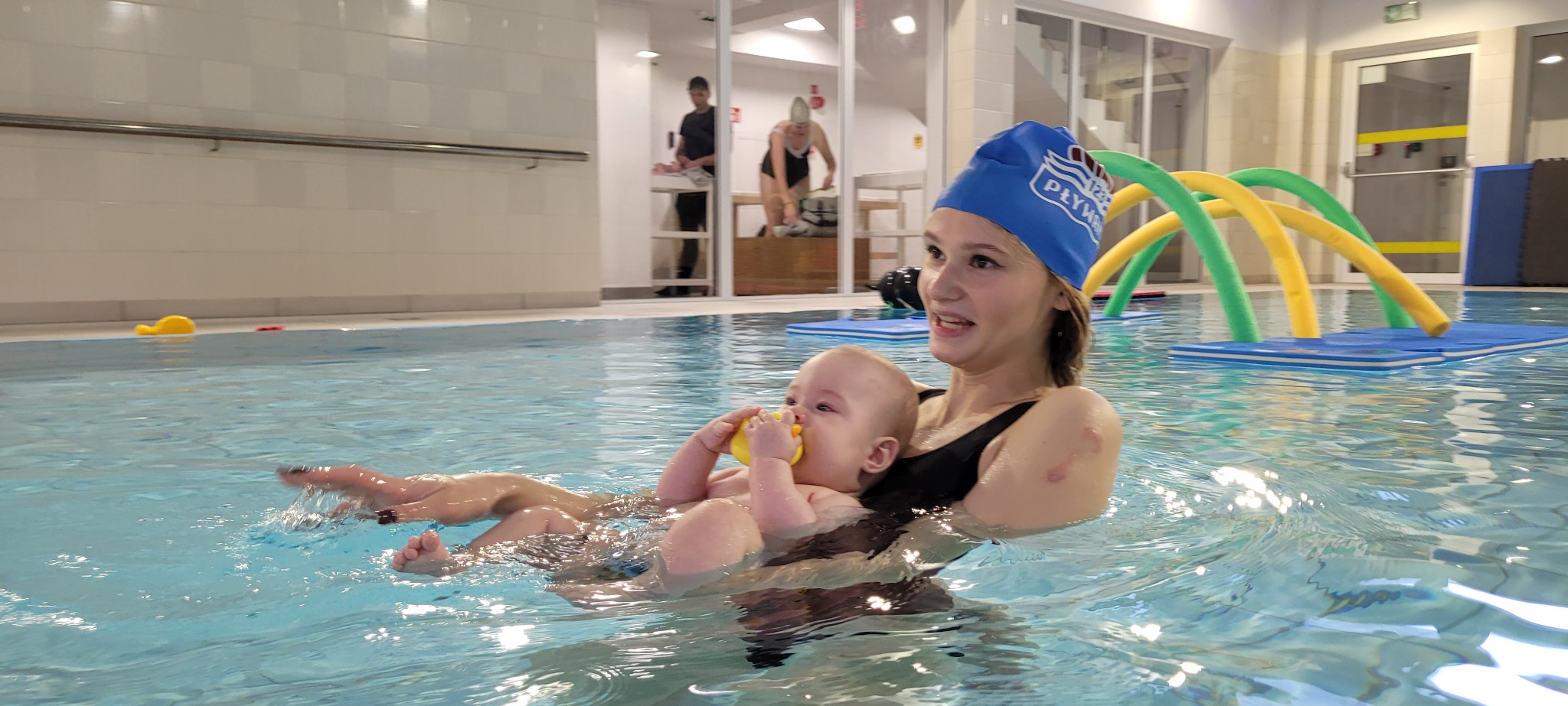 Instruktorka w czepku z logo, trzymająca niemowlę w wodzie, z żółtą zabawką w ustach. W tle basen z akcesoriami do pływania.