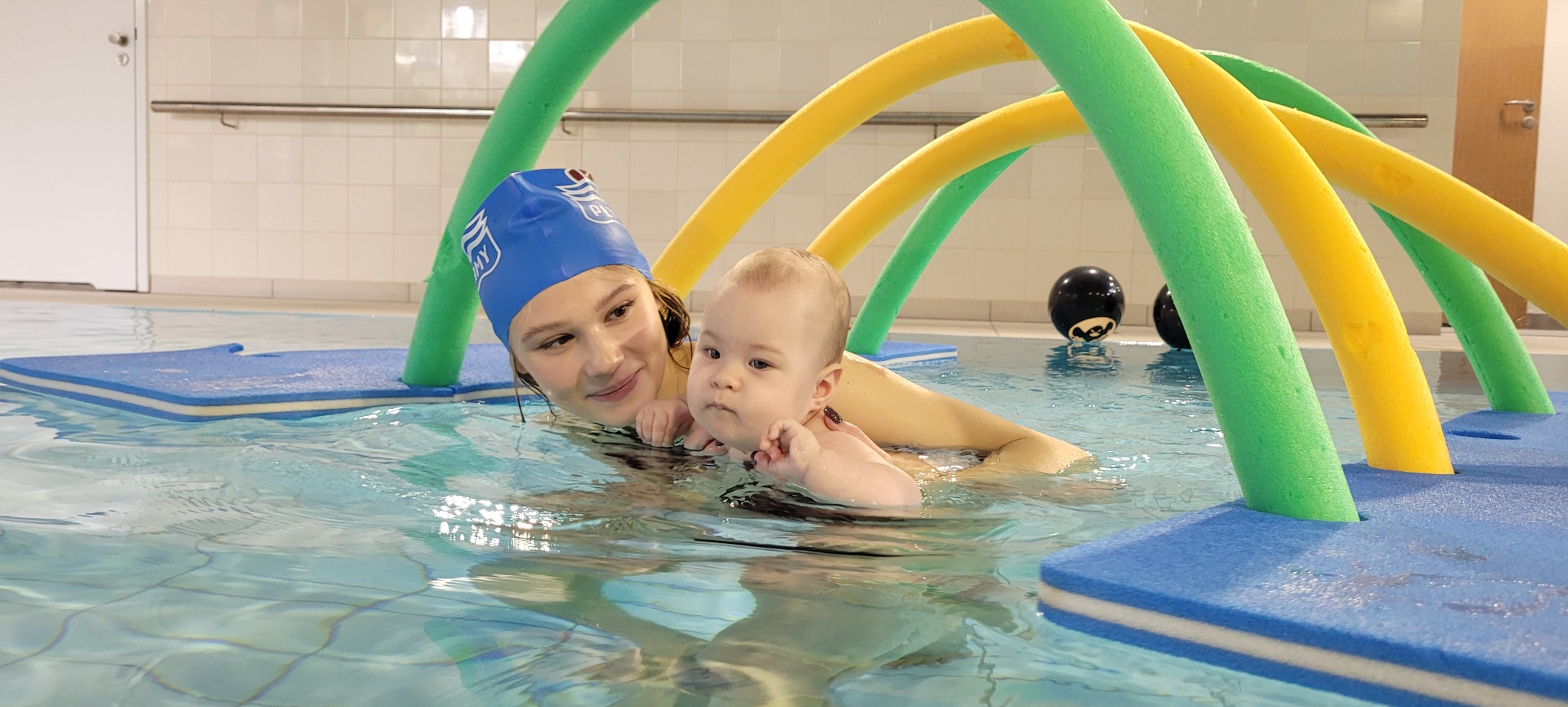 Instruktorka w czepku z niemowlęciem w basenie, otoczeni kolorowymi makaronami do pływania. Na wodzie unoszą się niebieskie piankowe maty.