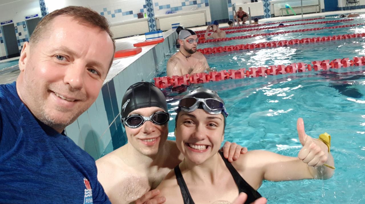 Uśmiechnięty instruktor z dwójką uczniów w czepkach i okularach pływackich w basenie, kciuk w górę, widoczny fragment logo na koszulce instruktora, niebiesko-białe płytki w tle.