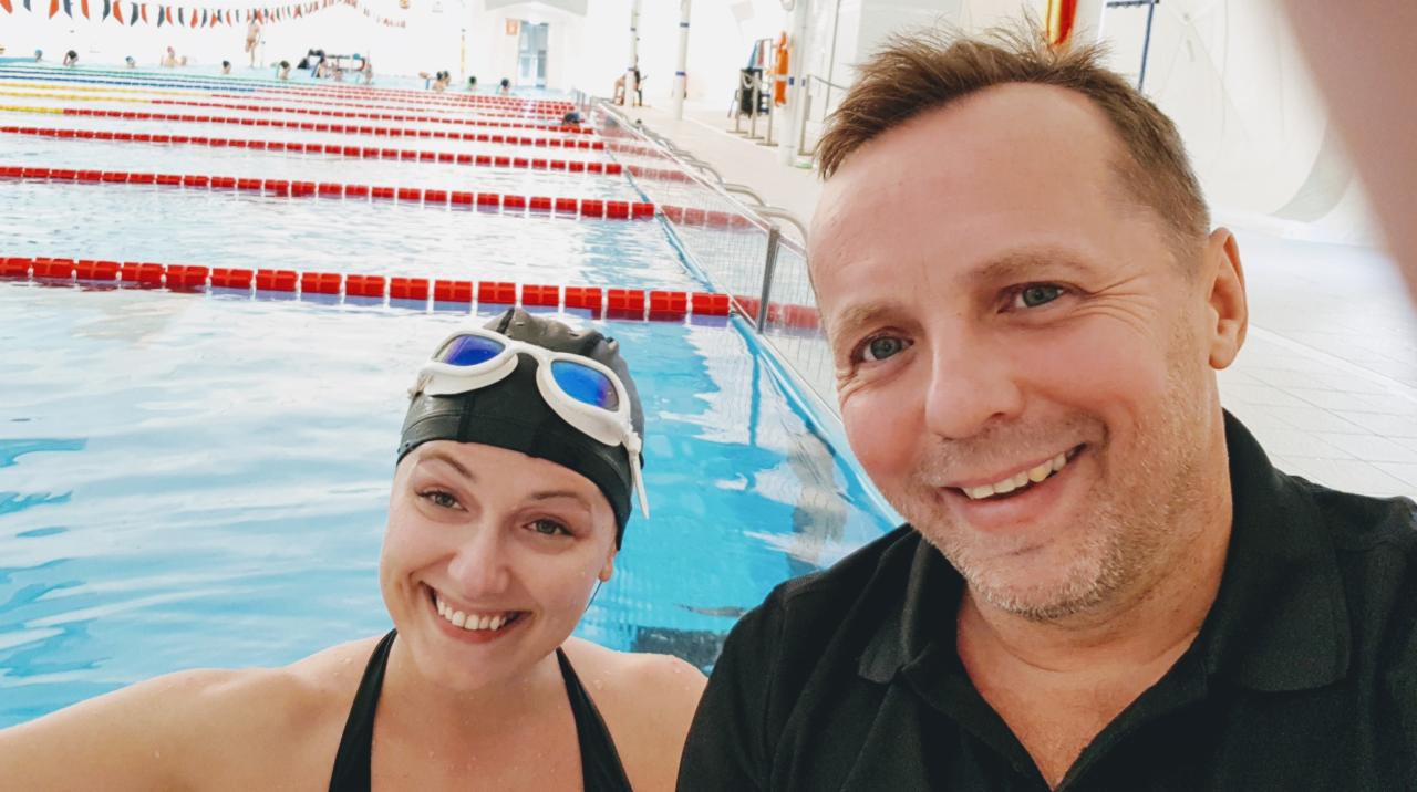 Uśmiechnięta kobieta w czepku i okularach pływackich oraz mężczyzna robią sobie selfie w basenie z torami.