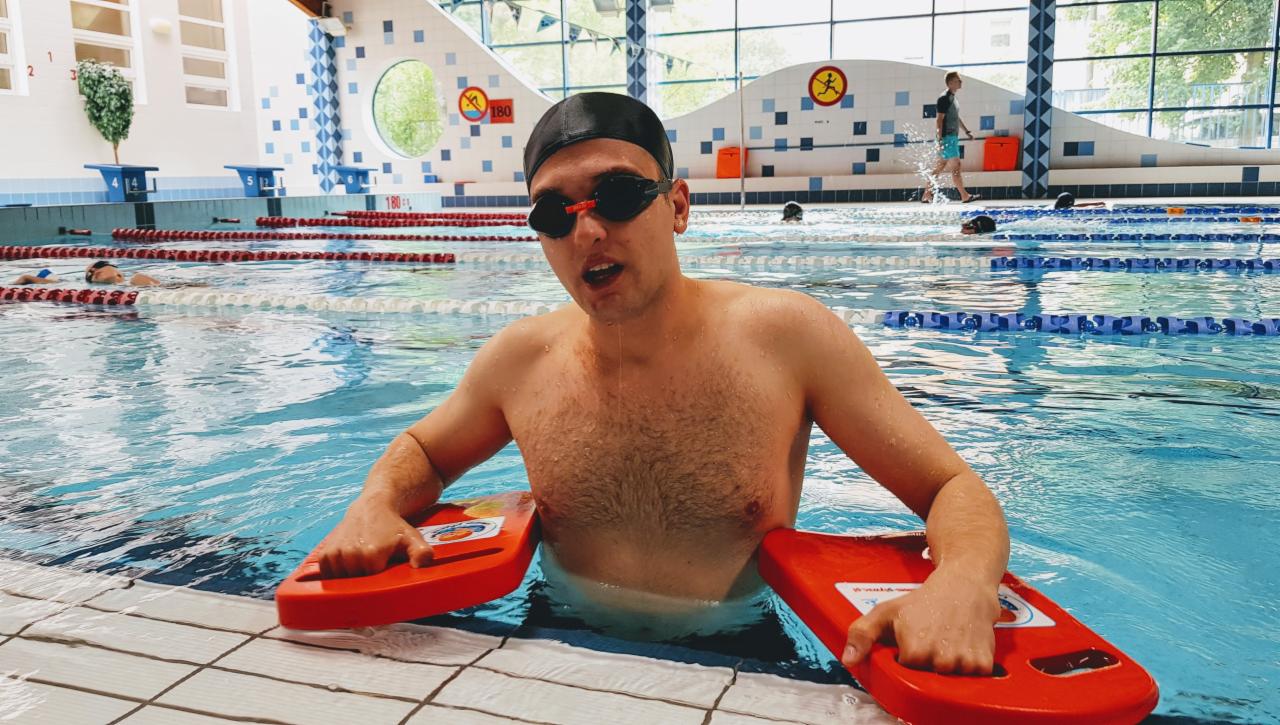 Młody mężczyzna w czepku i okularach pływackich, zanurzony w basenie, trzymający dwie czerwone deski do pływania, z otwartymi ustami i kroplami wody spływającymi po twarzy.
