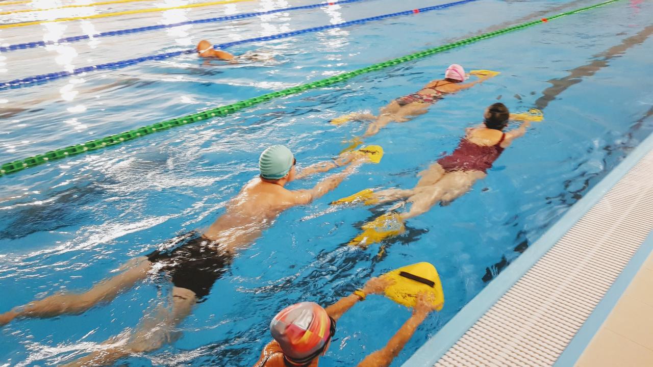 Kryty basen: grupa osób w różnym wieku ćwiczy pływanie z żółtymi deskami, widoczne tory oddzielone linami.