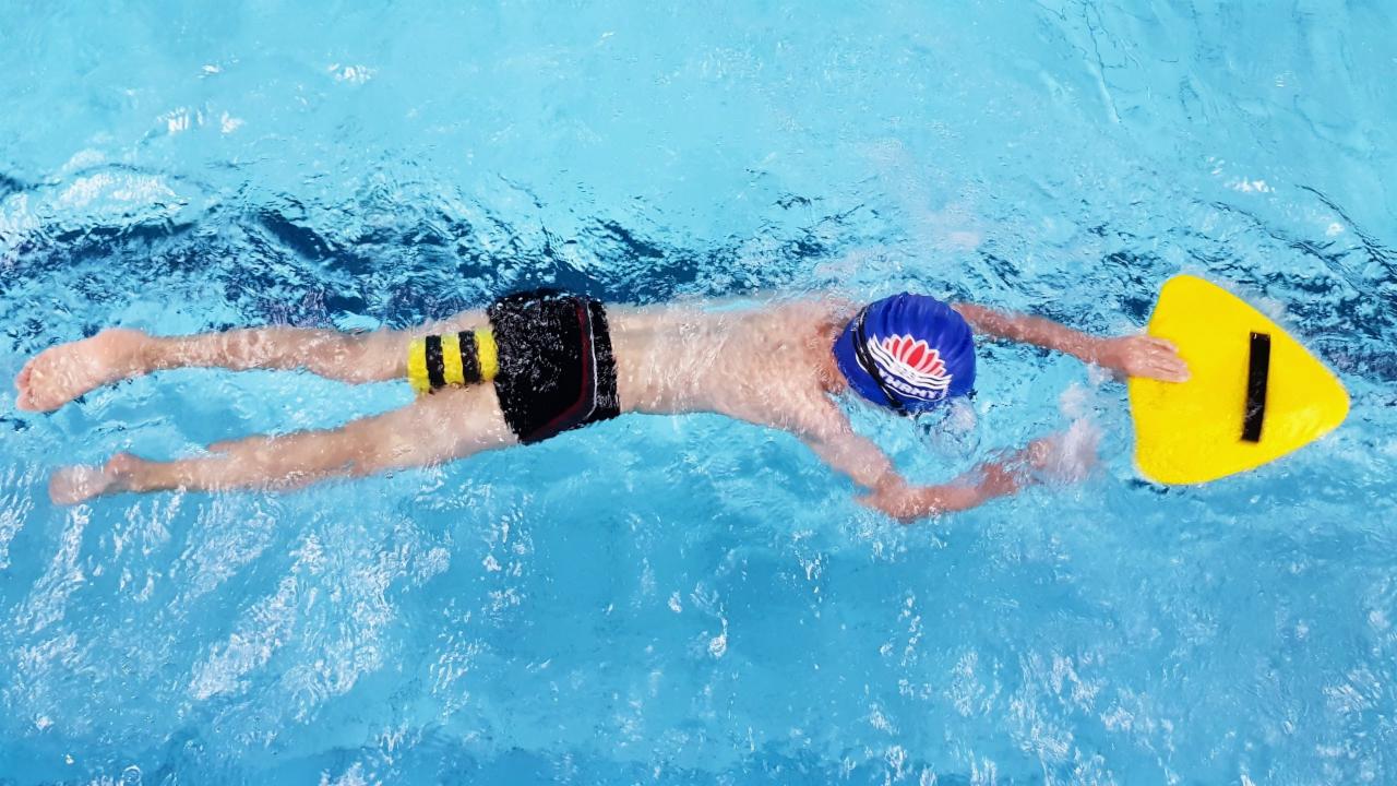 Chłopiec w czepku z nadrukiem, płynący na brzuchu w basenie z żółtą deską z czarnym paskiem, widok z góry.