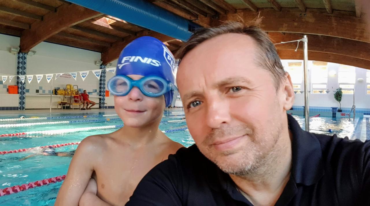 Selfie instruktora z uśmiechniętym chłopcem w czepku i okularach pływackich na tle basenu z widocznym ratownikiem