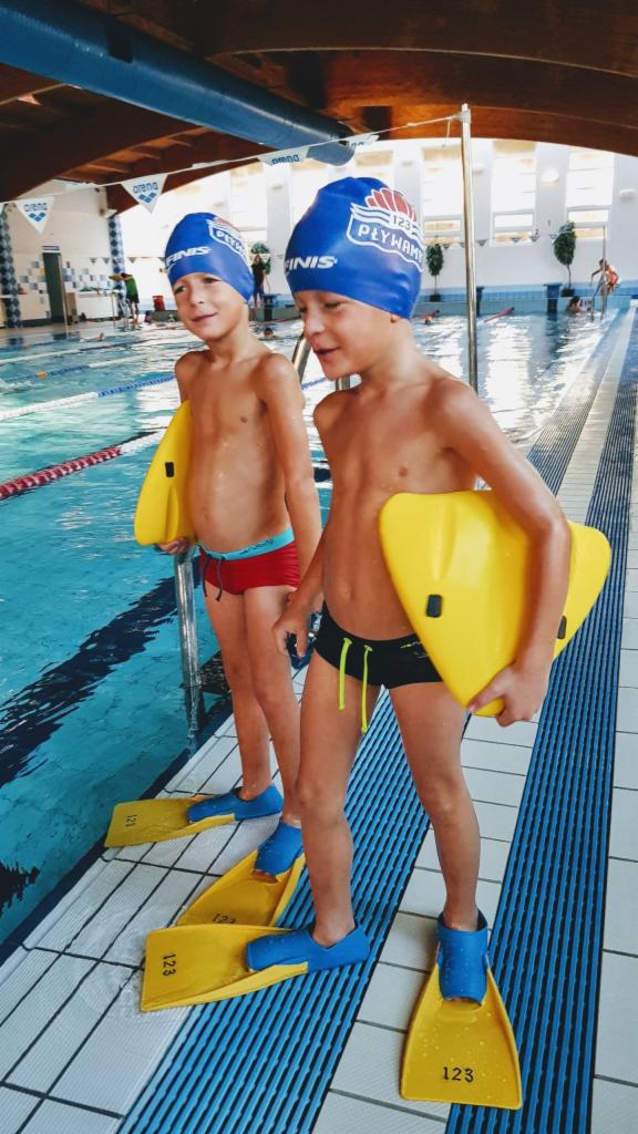 Dwóch chłopców w czepkach i płetwach, trzymających deski do pływania, stoi na brzegu basenu w Warszawie, gotowych do zajęć.