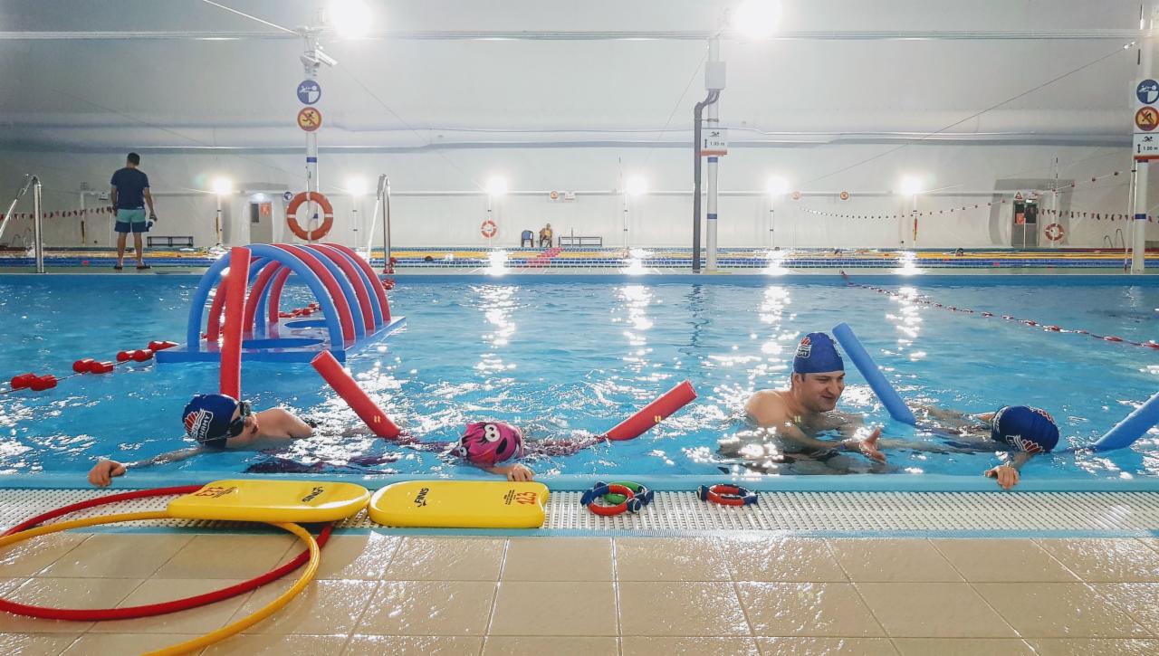 Instruktor pływania z uśmiechem pomaga dzieciom w nauce pływania w basenie, używając piankowych rurek i desek.