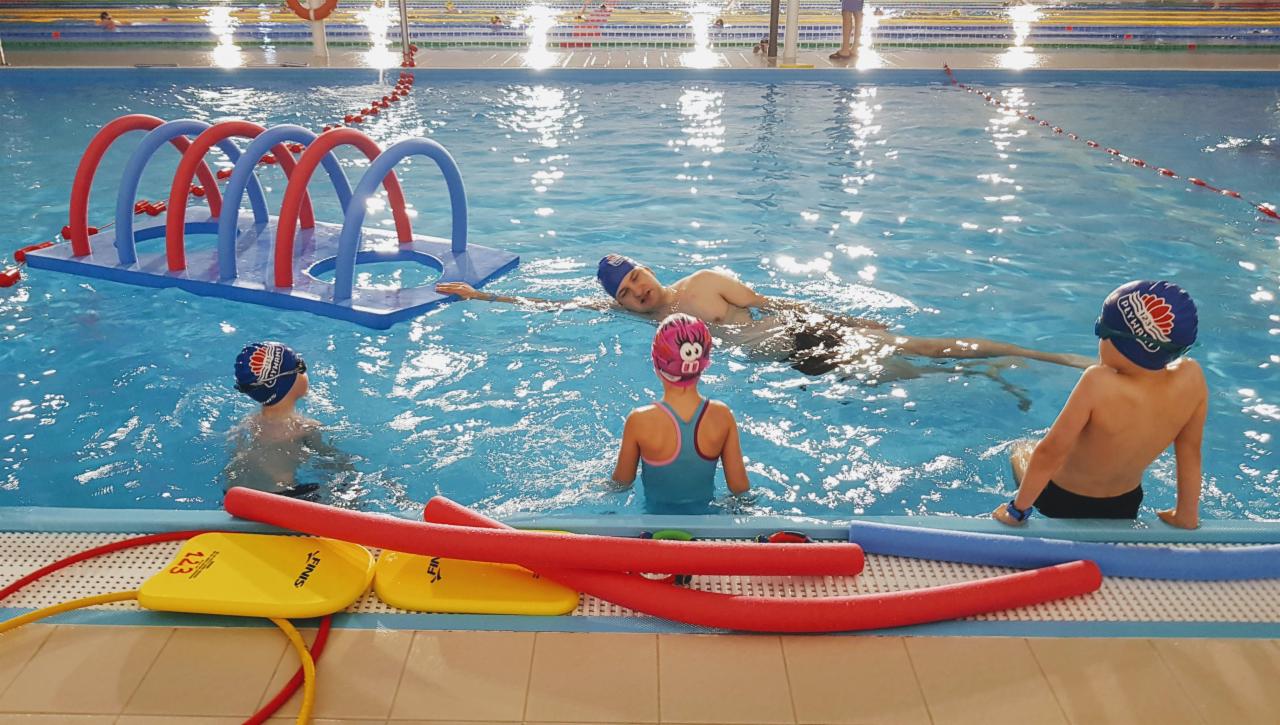 Kryty basen z osobami w trakcie zajęć: instruktor pomaga dziecku płynąć, w tle pływająca konstrukcja z rur. Widoczne deski do pływania i piankowe wałki.