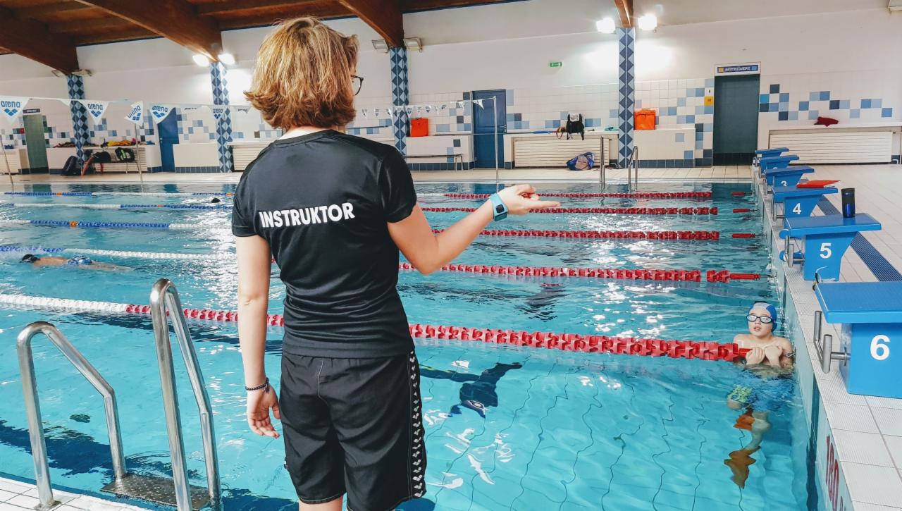 Instruktorka w czarnej koszulce z napisem 'INSTRUKTOR' na plecach, stojąca przy basenie z kilkoma pływającymi osobami; widoczny chłopiec w wodzie przy brzegu, niebieskie słupki startowe z numerami.