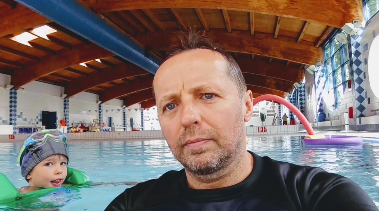 Selfie instruktora pływania z dzieckiem w basenie; dziecko w czepku i okularach pływackich oraz z zielonymi motylkami na ramionach, w tle widoczne akcesoria do pływania.