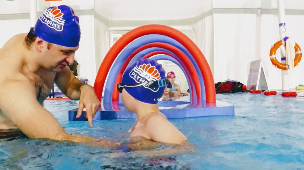 Instruktor w basenie pomaga dziecku w czepku z napisem 'Pływamy' i goglami; w tle widoczne kolorowe tunele z pianki i inne dzieci.