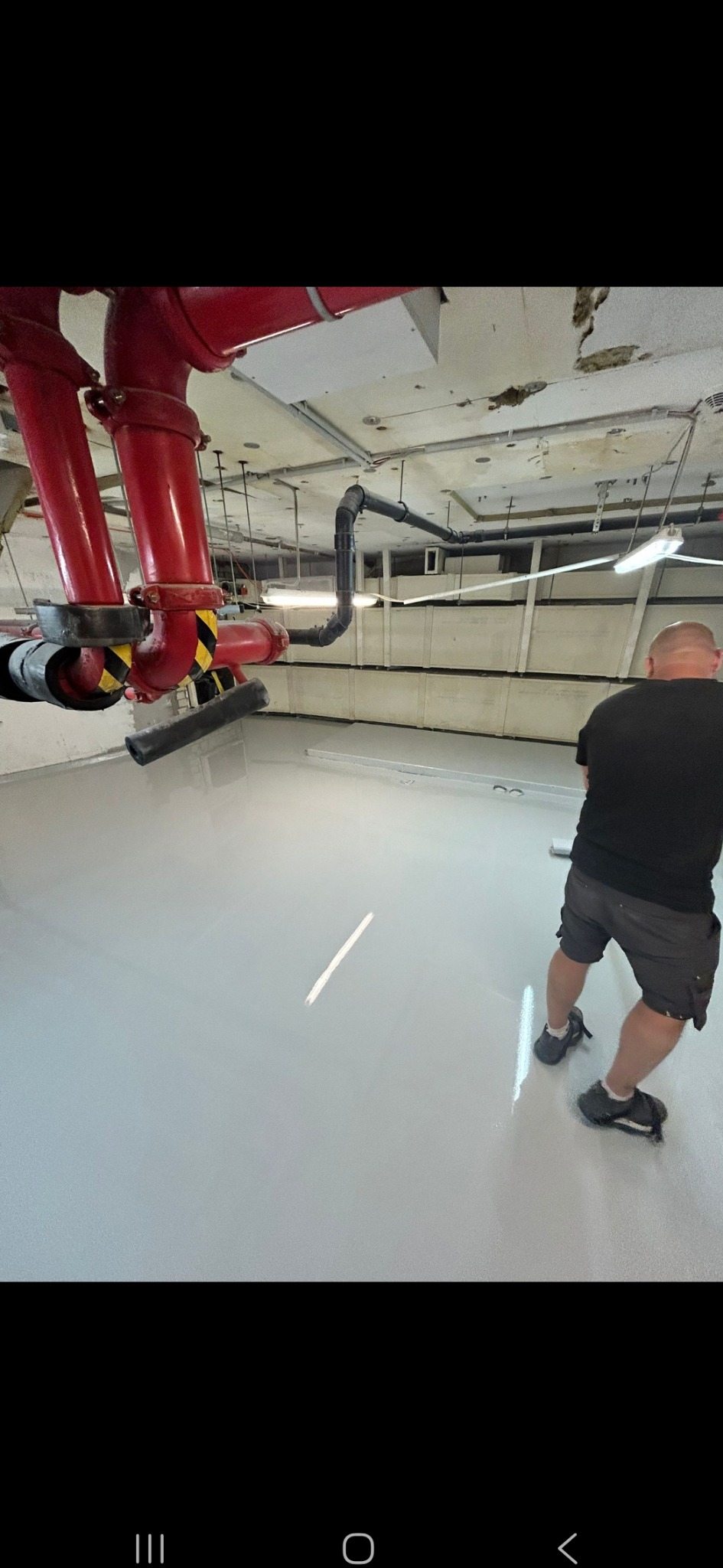 Jasnoszara, gładka posadzka w hali z czerwonymi rurami pod sufitem. Mężczyzna w szortach rozprowadza masę szpachlową. Widoczne ślady po wałku.