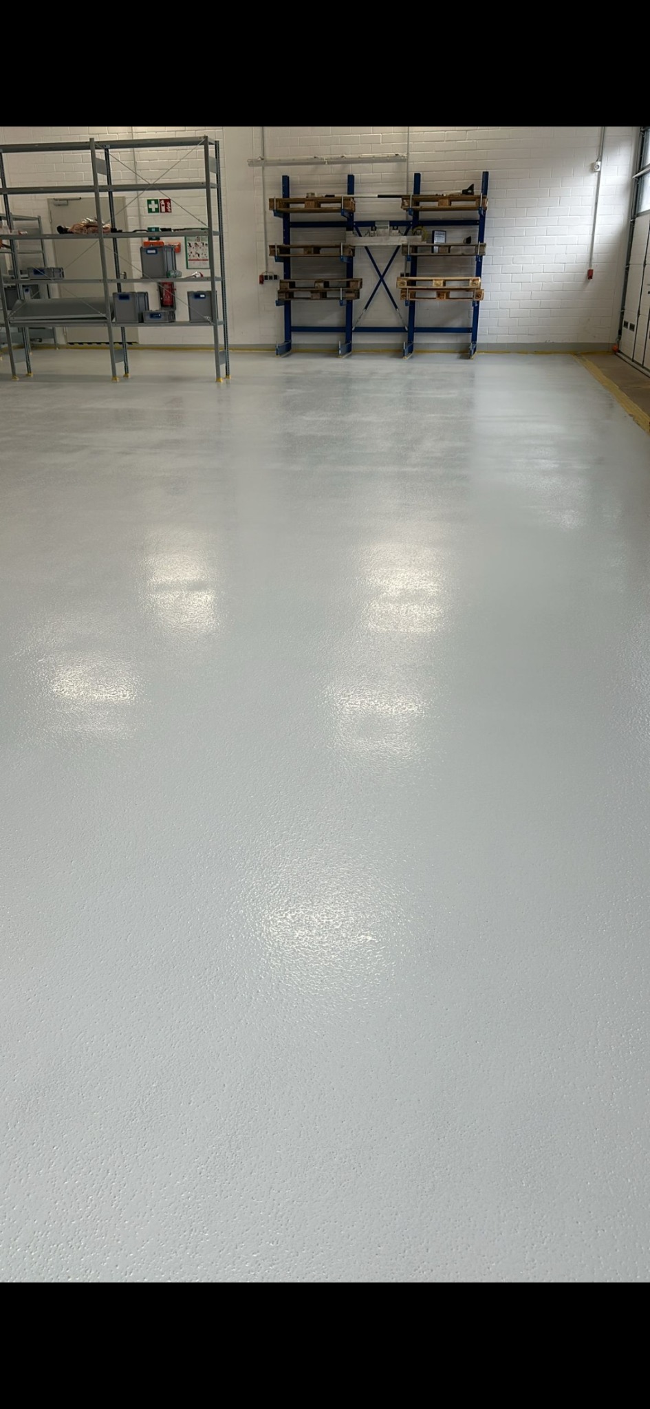 Biała, gładka posadzka w hali magazynowej, z metalowymi regałami i ceglaną ścianą w tle. Widoczne ślady po malowaniu na podłodze.