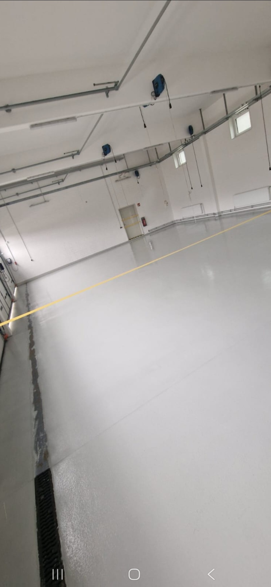 Biała, przemysłowa posadzka żywiczna w hali produkcyjnej, z widocznymi instalacjami pod sufitem i żółtą taśmą wyznaczającą strefę. Gładka powierzchnia, jasne oświetlenie.