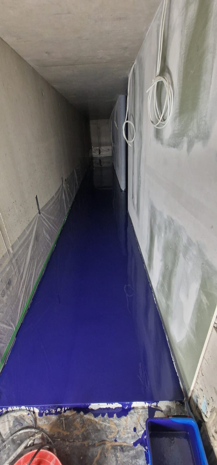 Niebieska, błyszcząca posadzka w wąskim pomieszczeniu w trakcie prac wykończeniowych. Ściany przygotowane do dalszych prac, widoczne kable i narzędzia malarskie.