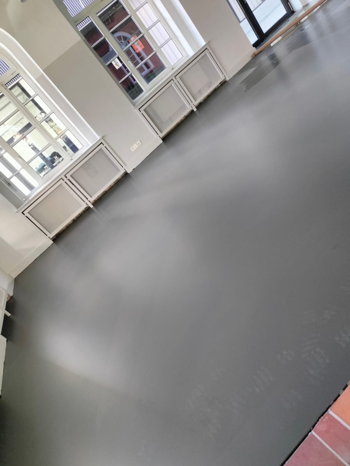 Szara, świeżo pomalowana podłoga w jasnym pomieszczeniu z oknami i grzejnikami. Widoczne ślady stóp na powierzchni. Ujęcie z perspektywy pod kątem.