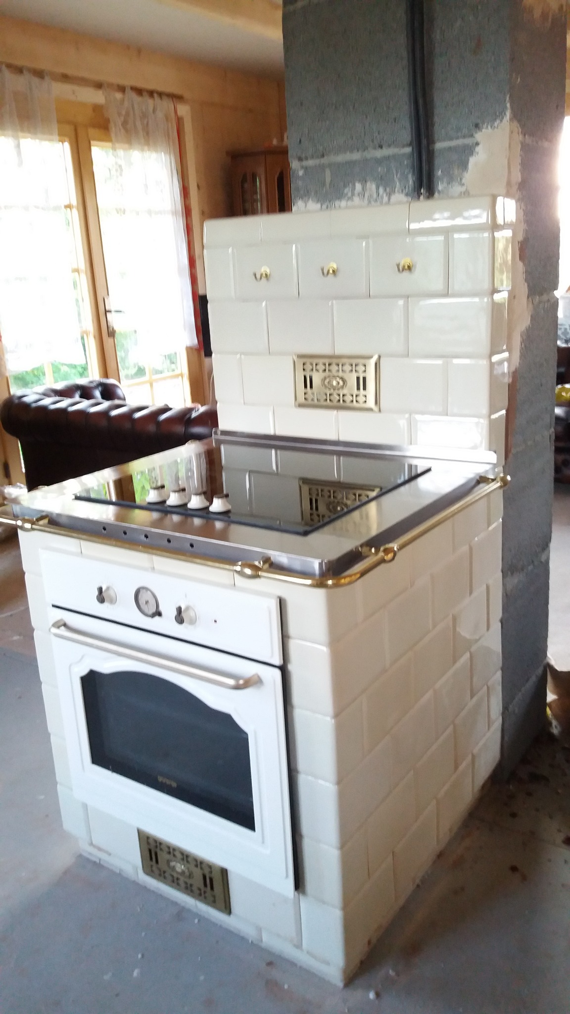 Biały piec kaflowy z piekarnikiem i płytą grzewczą, wykończony złotymi detalami, w surowym wnętrzu drewnianego domu.