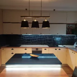 Jasna kuchnia z wyspą i czarnym blatem, oświetlona trzema wiszącymi lampami. Czarny mozaikowy backsplash z podświetleniem LED. Na wyspie dekoracyjna misa.