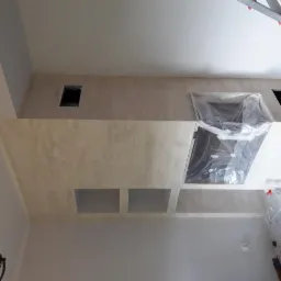 Ściana z nałożoną strukturą dekoracyjną, widoczne otwory wentylacyjne i zabezpieczony folią kominek, drabina w tle.