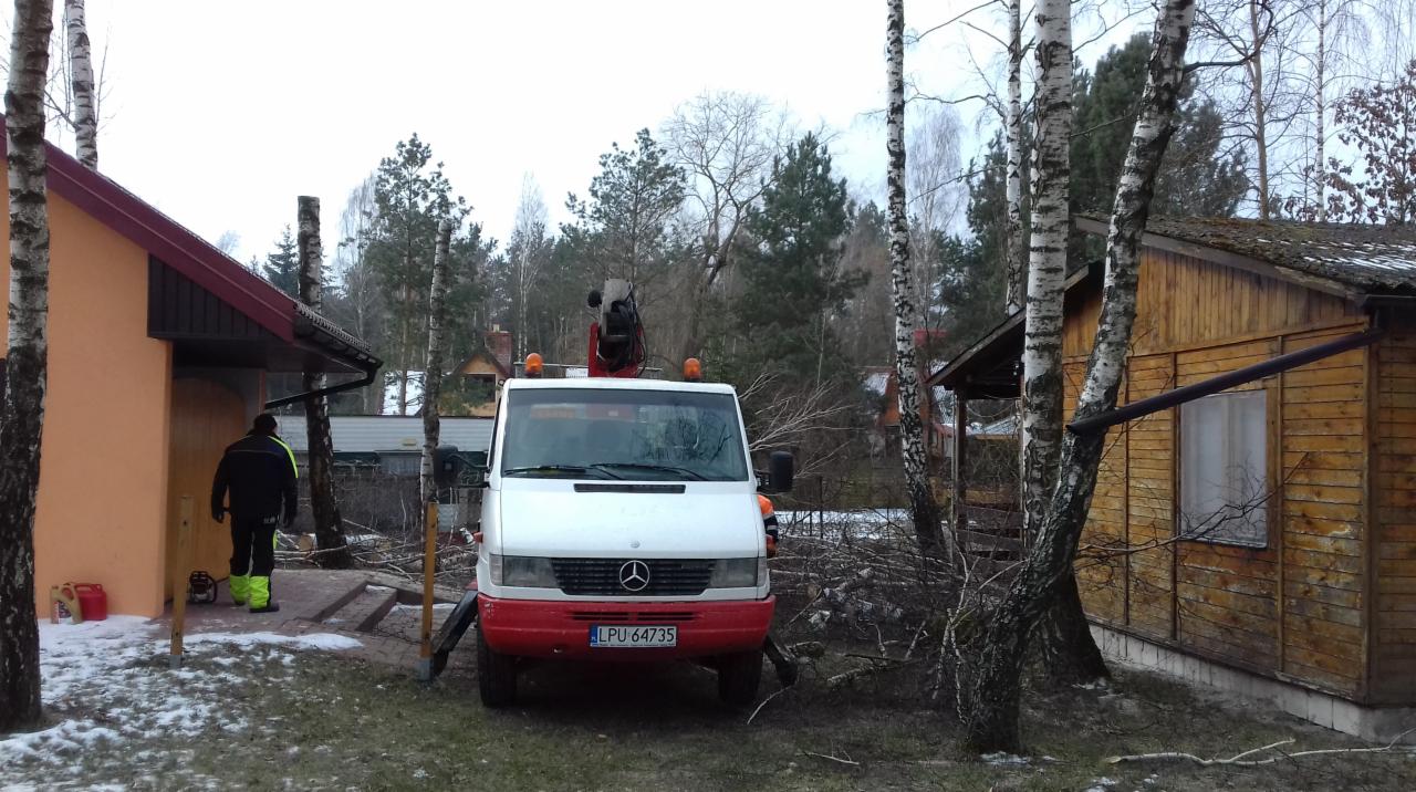 Biały samochód z podnośnikiem na tle dwóch domków i powalonych gałęzi, pracownik w odblaskowym ubraniu w tle, pochmurny dzień.