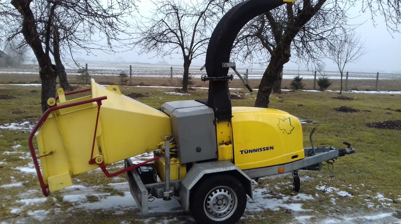 Żółta, mobilna rębarka do gałęzi marki Tünnissen na trawiastym terenie z resztkami śniegu, drzewa bez liści w tle.
