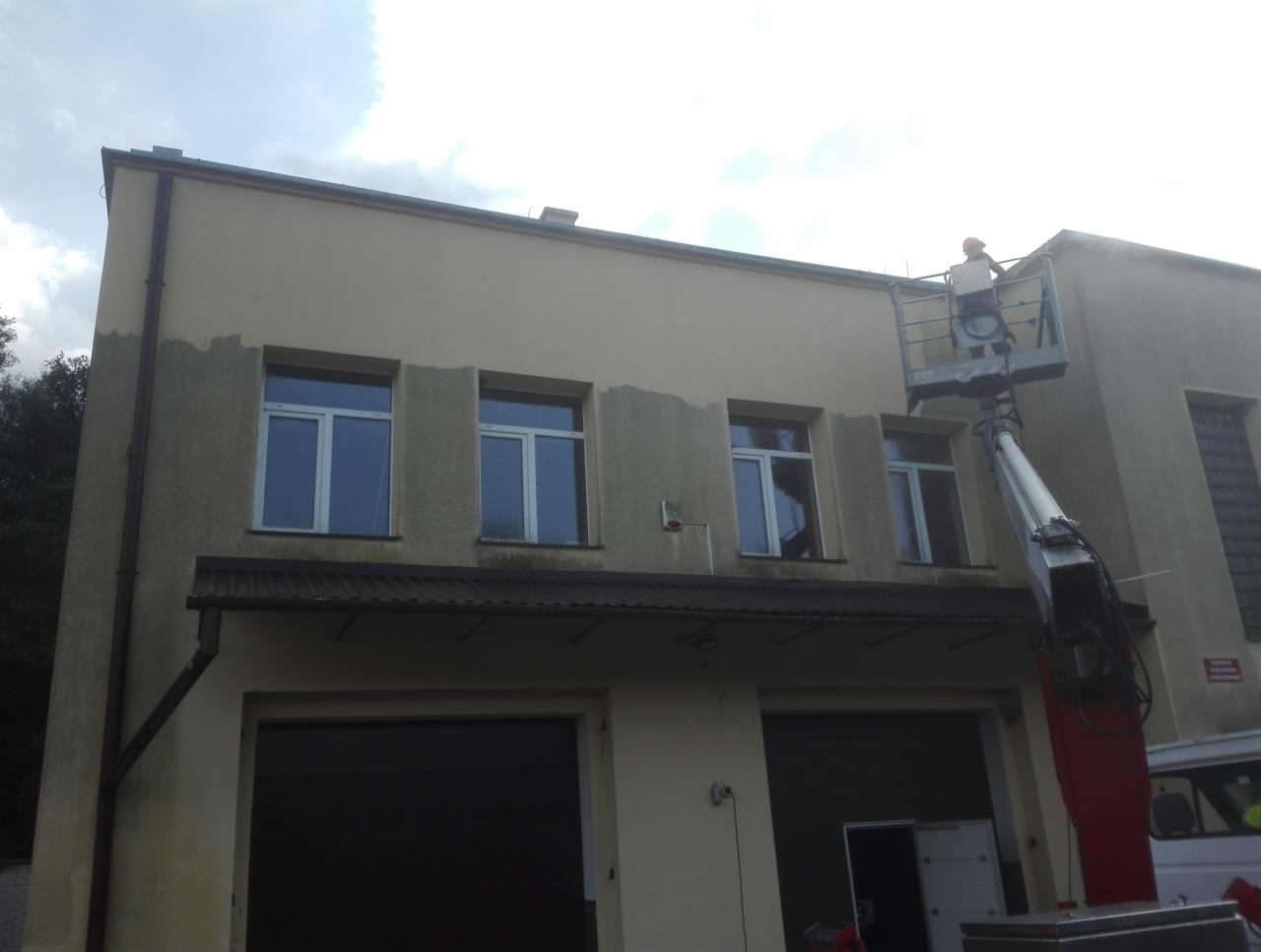 Pracownik w pomarańczowym kasku na podnośniku koszowym maluje elewację budynku na beżowo, widoczne fragmenty surowej ściany i pomalowanej powierzchni.