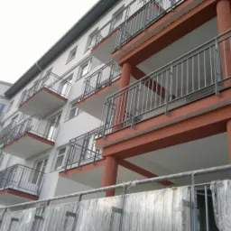 Nowoczesny budynek wielorodzinny z balkonami zabezpieczonymi balustradami nierdzewnymi, widok z dołu, z fragmentem zabezpieczenia budowy na pierwszym planie.