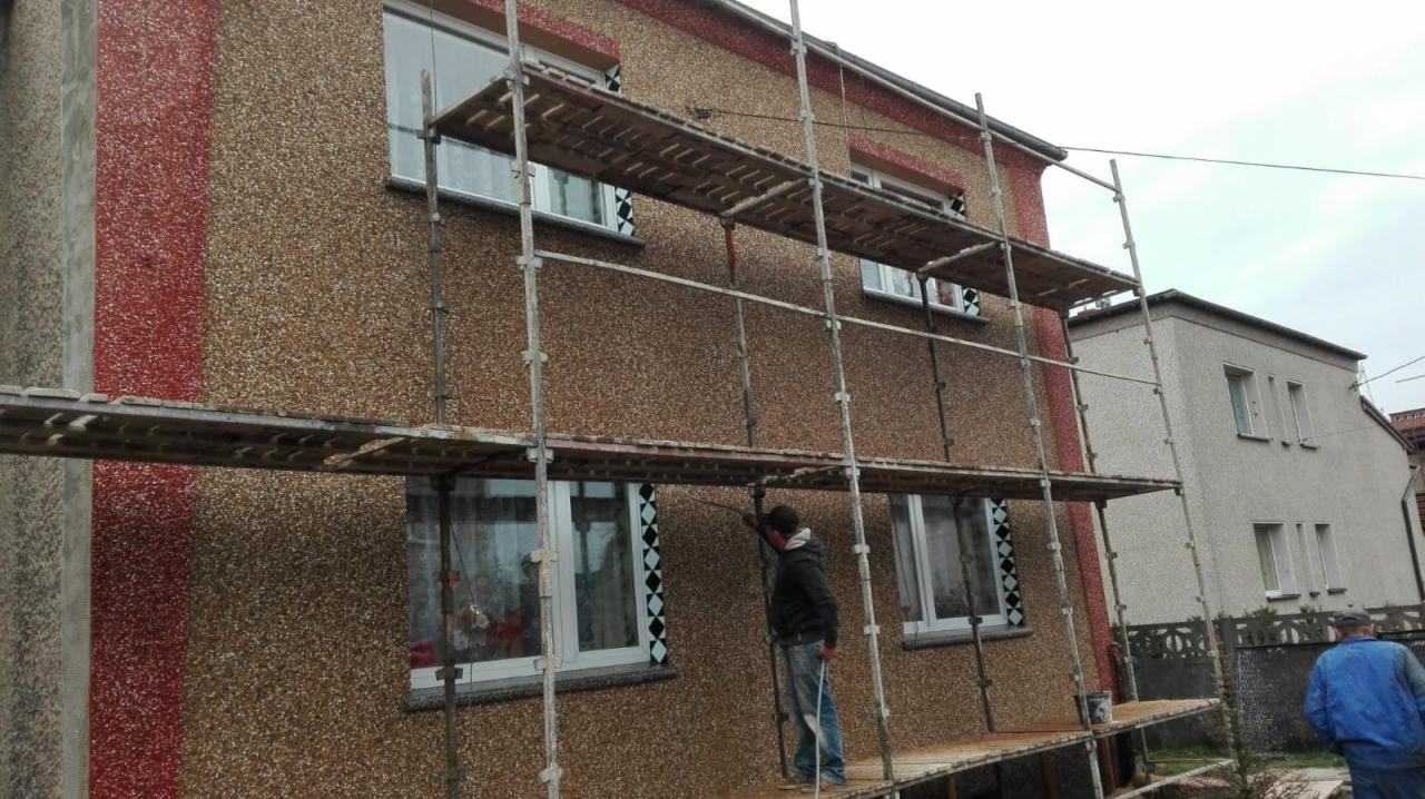 Dwukondygnacyjny dom z elewacją typu baranek w odcieniach brązu i czerwieni, częściowo zasłonięty rusztowaniem, na którym stoi pracownik w trakcie wykonywania prac elewacyjnych.