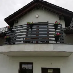 Balkon z poziomą, ciemnoszarą konstrukcją zabezpieczającą, udekorowany doniczkami z różowymi kwiatami, widok z dołu na tle szarego nieba i fragmentu dachu.
