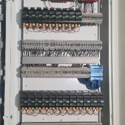 Otwarta, biała szafa sterownicza z widocznymi modułami Loxone, licznymi złączkami, przewodami o różnych kolorach i oznaczeniami, zamontowana na tle surowego stropu.