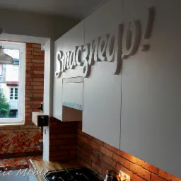 Białe szafki kuchenne z napisem 'Smacznego!' na tle ceglanej ściany i okna z widokiem na zewnątrz, z fragmentem blatu i płyty grzewczej w dolnej części kadru.