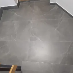 Szare płytki podłogowe imitujące marmur, ułożone w łazience z ciemnoszarą listwą przypodłogową i fragmentem drabiny malarskiej w rogu.