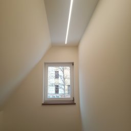 Firma REMONTOWO-BUDOWLANA Piotr Soltys - Jasne wnętrze poddasza z oknem i liniowym oświetleniem LED. Ściany w odcieniach beżu, drewniana podłoga. Widok z okna na budynek i drzewo.