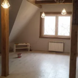 Poddasze z drewnianymi belkami, jasną podłogą, białymi ścianami, oknem z grzejnikiem i trzema nowoczesnymi lampami wiszącymi, widoczny pojemnik z farbą i biały stolik