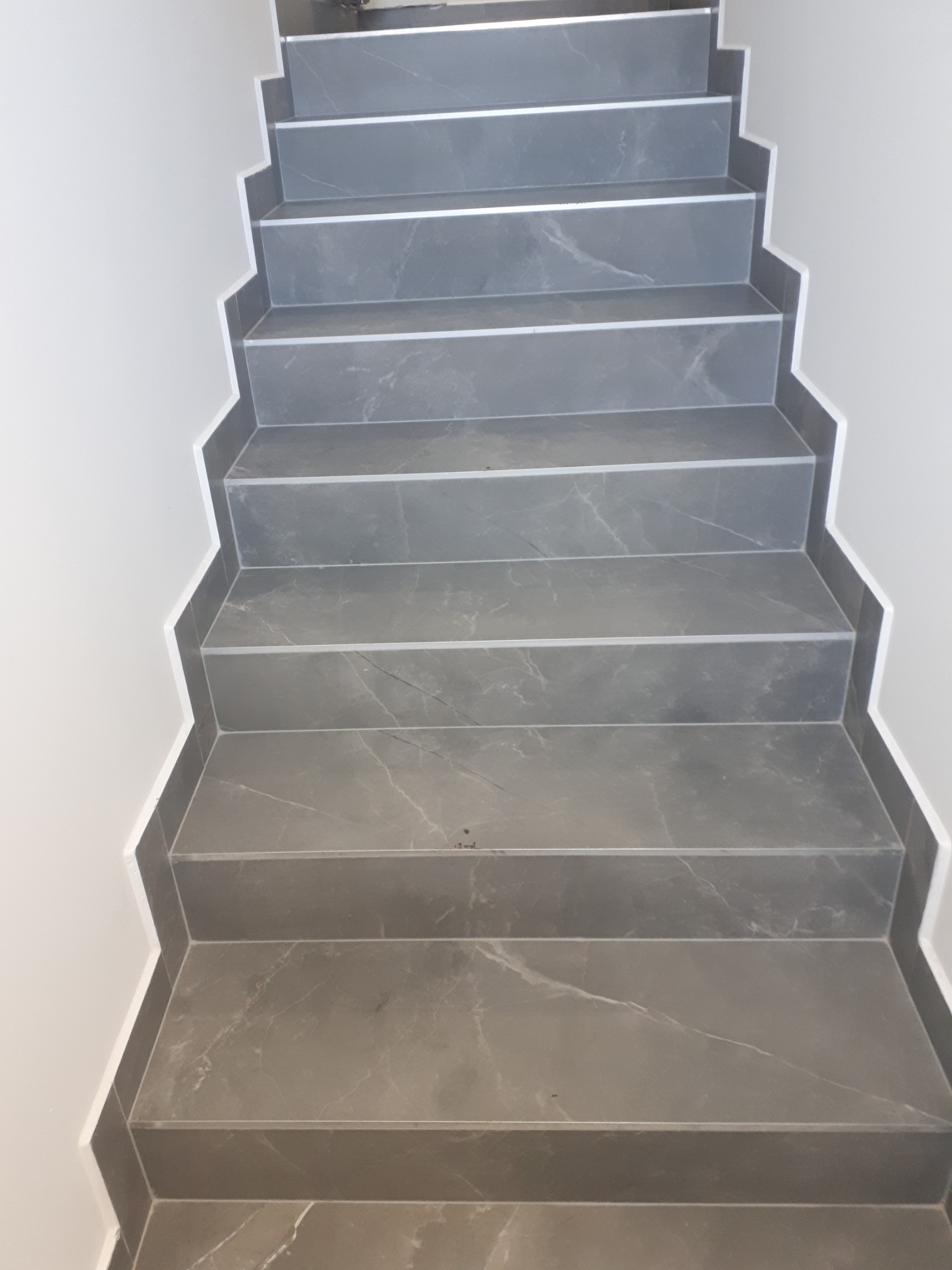 Wykończone schody wewnętrzne wyłożone szarymi płytkami imitującymi marmur, z białymi listwami wykończeniowymi przy ścianie.