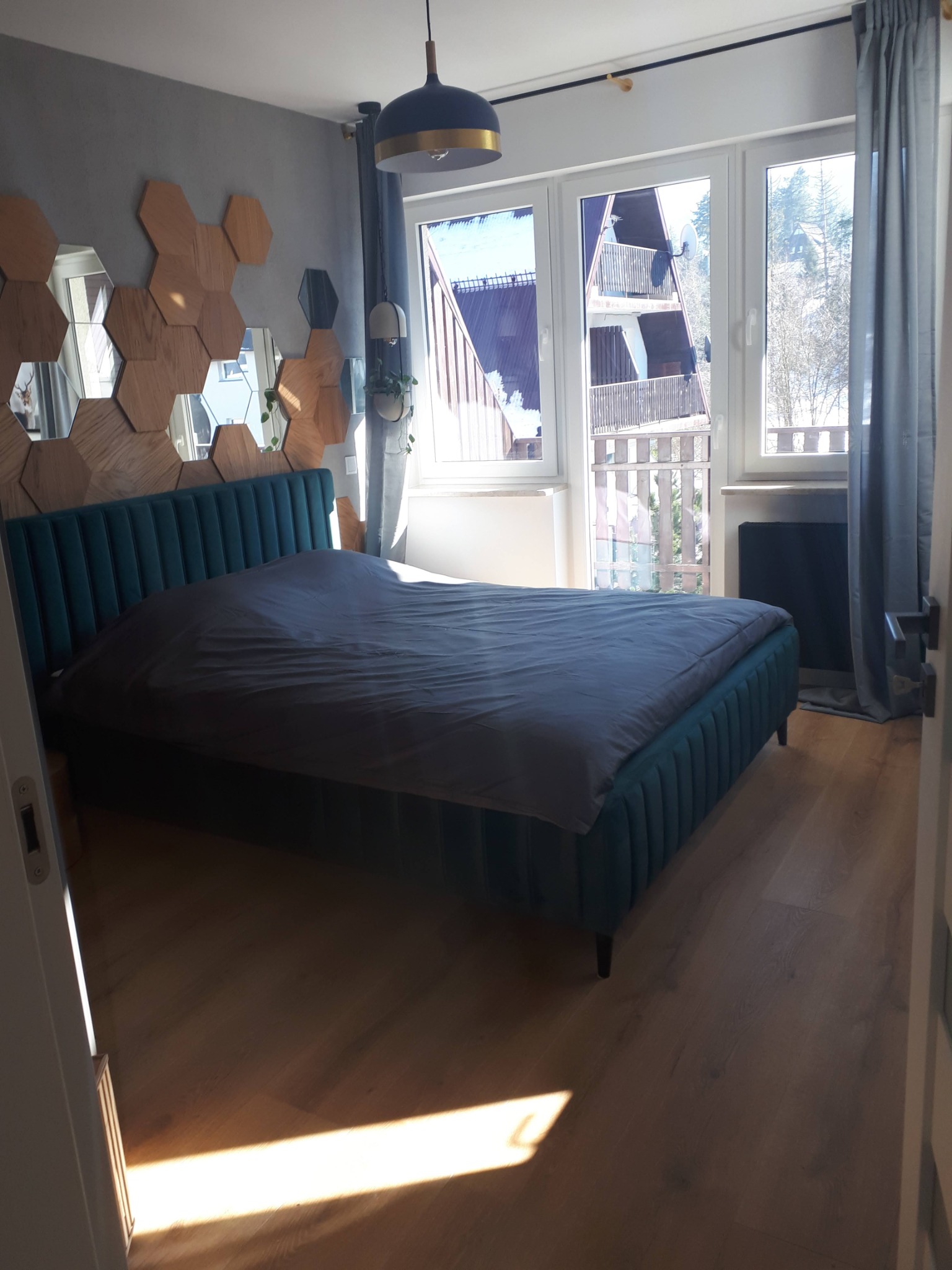 Sypialnia z turkusowym łóżkiem i drewnianą ścianą w kształcie plastra miodu, widok z otwartych drzwi na parkiet z promieniem słońca.