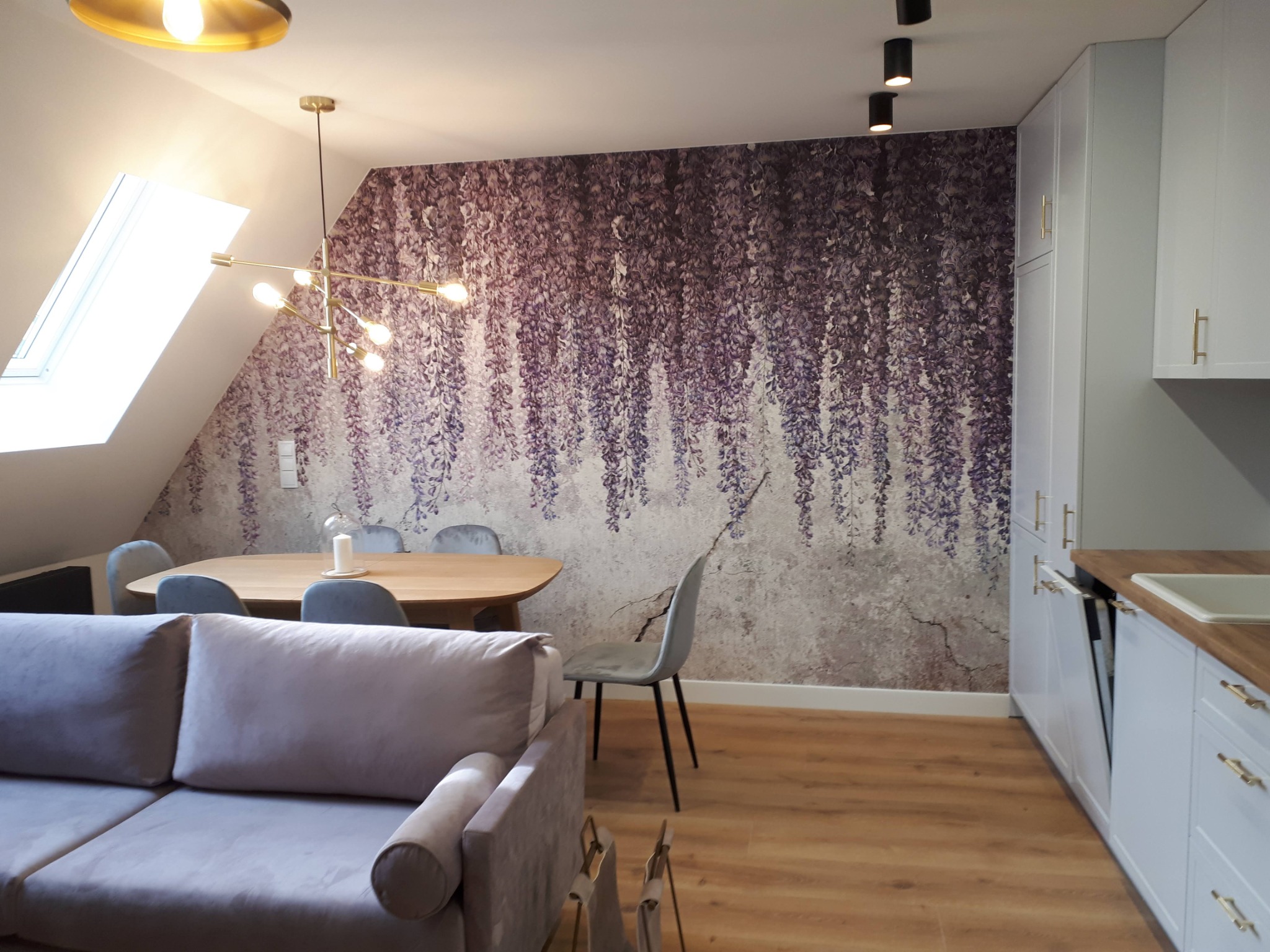Jasne wnętrze mieszkania na poddaszu z fioletową tapetą w motywy kwiatowe, drewnianym stołem i szarymi krzesłami, oświetlone nowoczesną lampą wiszącą i reflektorami sufitowymi, z widoczną częścią...