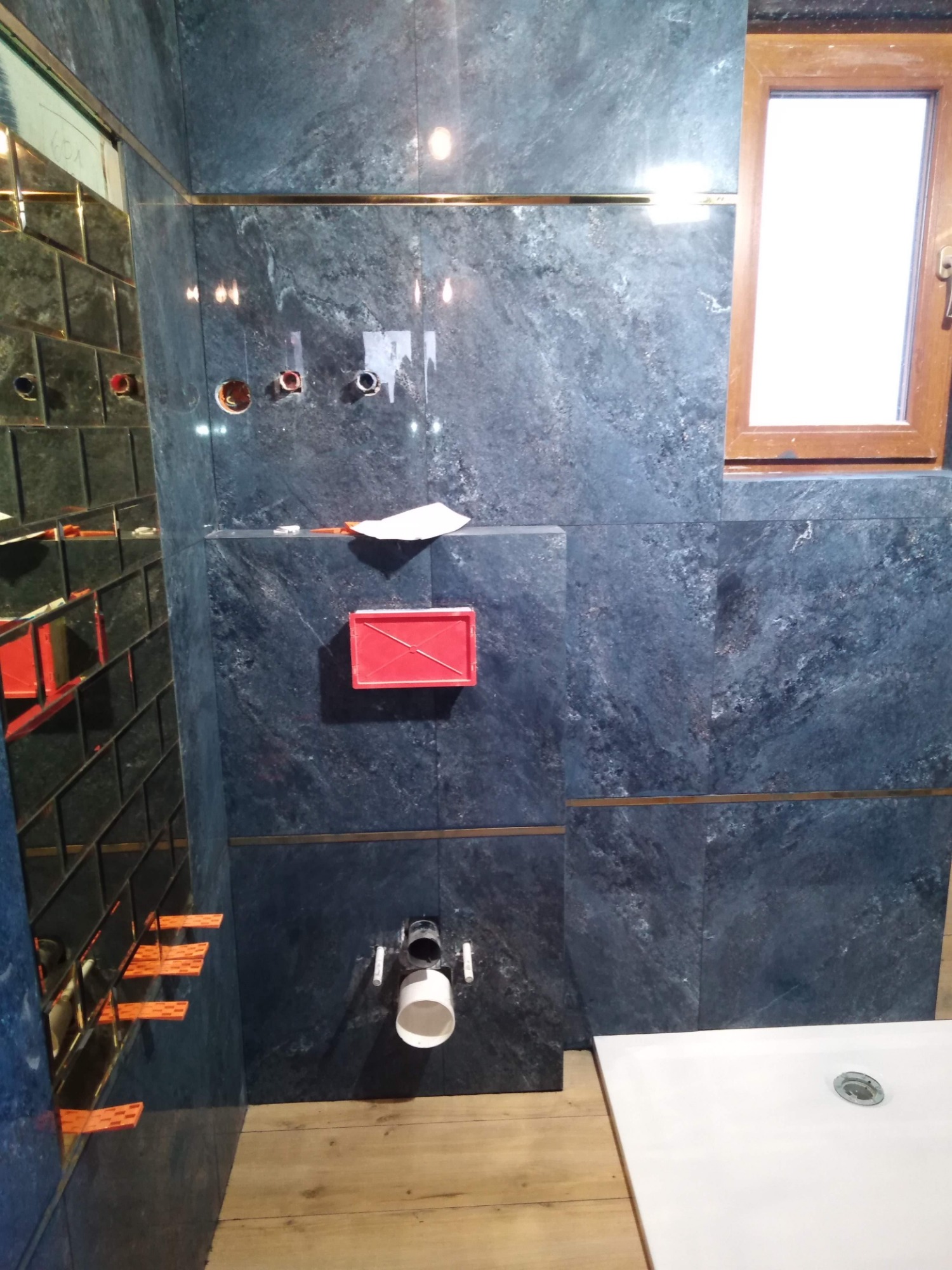 Montaż ciemnych, marmurkowych płytek w łazience, widoczne otwory pod armaturę, fragment okna z drewnianą ramą, odpływ liniowy i przymocowane poziomicą płytki lustrzane.