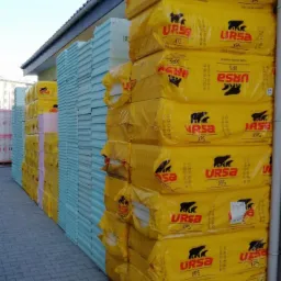 Stosy zapakowanych płyt izolacyjnych URSA XPS w żółtych foliach, obok układanka niebieskich płyt styropianowych, składowane na zewnątrz budynku.