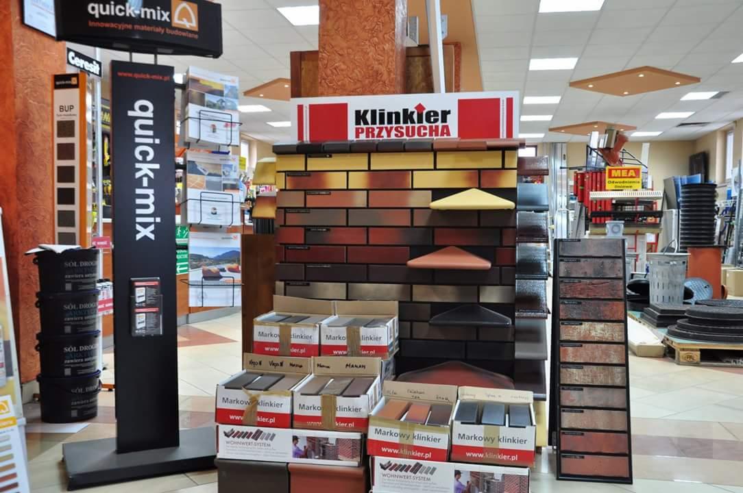 Ekspozycja klinkieru w sklepie budowlanym: próbki cegieł w różnych odcieniach, ułożone na ścianie, oraz pudełka z cegłami z logo producenta, oświetlone światłem z sufitu.