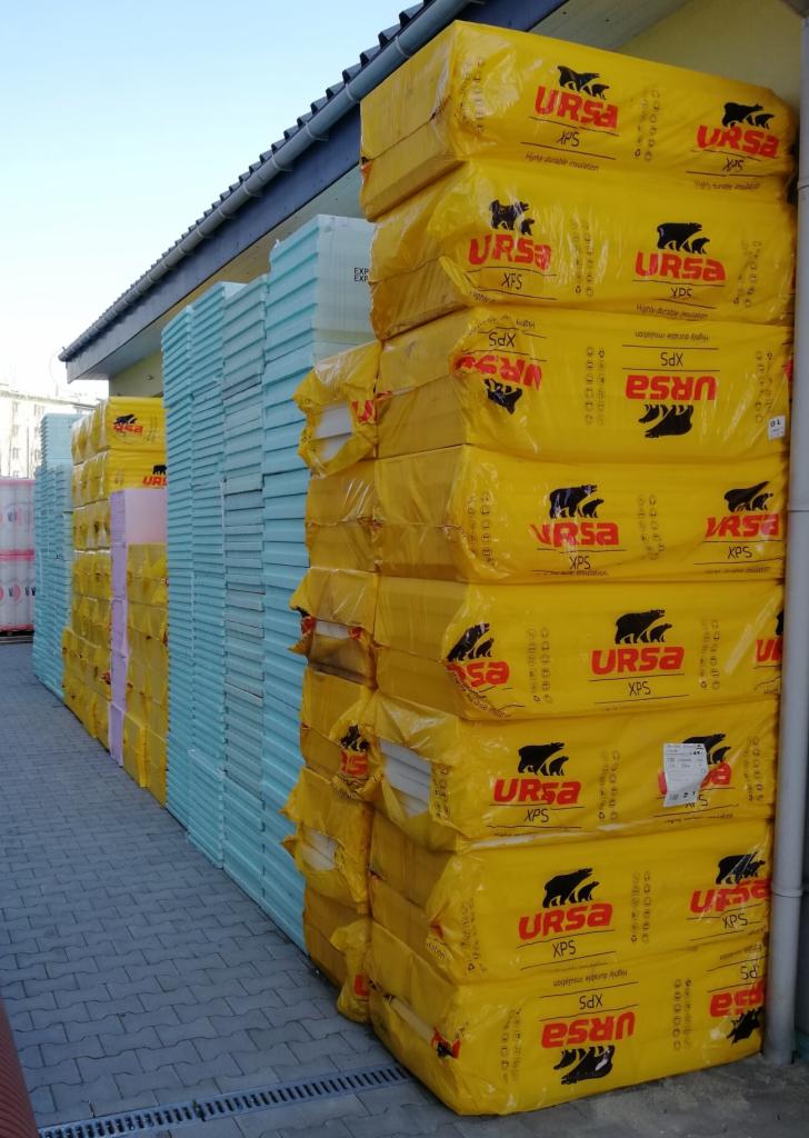 Stosy zapakowanych płyt izolacyjnych URSA XPS w żółtych foliach, obok układanka niebieskich płyt styropianowych, składowane na zewnątrz budynku.