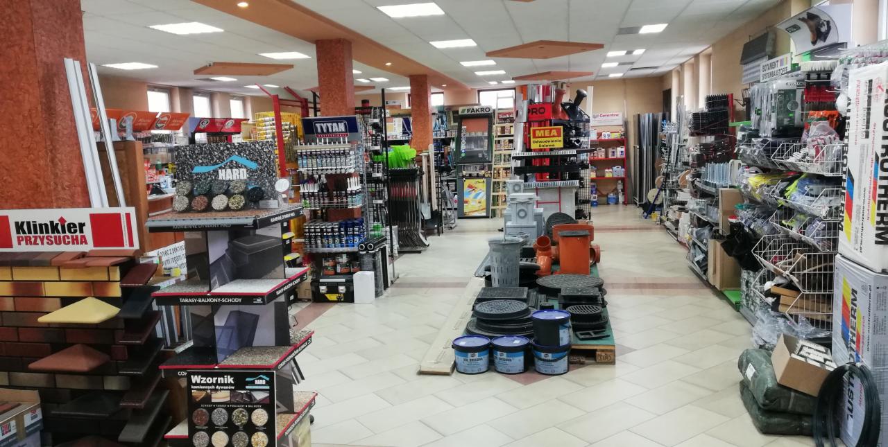 Wnętrze sklepu budowlanego z ekspozycją materiałów wykończeniowych, w tym klinkieru, kostki brukowej i systemów odwadniających, z widocznymi regałami pełnymi farb, narzędzi i akcesoriów.